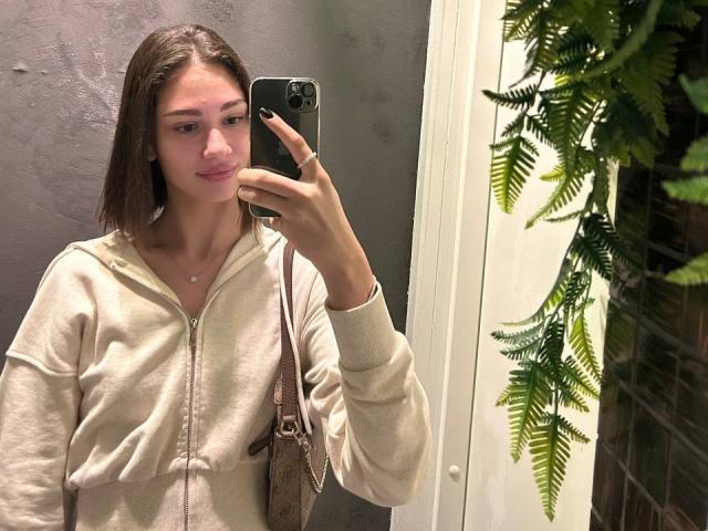 TobiQuinby mit glattem, braunem Haar, rosafarbenen Lippen, trägt eine beige Kapuzenjacke mit Reißverschluss und macht ein Selfie mit einem grünen Handy. Sie sieht süß aus, trägt eine kleine, weiße Halskette, im Hintergrund sind Farne zu sehen..