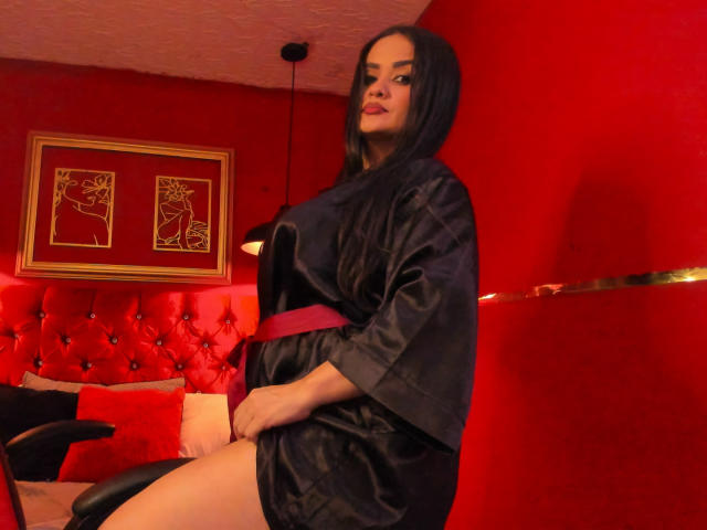 AnissaAli in a black satin robe, pink belt, sitting with her hand on her thigh, red room, tufted red wall, black chair, red pillow, black hair, red lipstick.