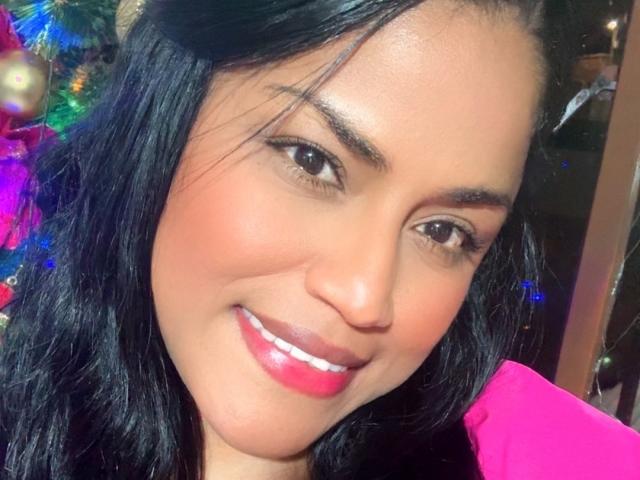 IvannaDuran69, con piel de tono marrón medio, cabello negro ondulado, ojos marrones, lápiz labial rojo y una ligera sonrisa, vestida con una blusa rosa, luciendo linda y atractiva..