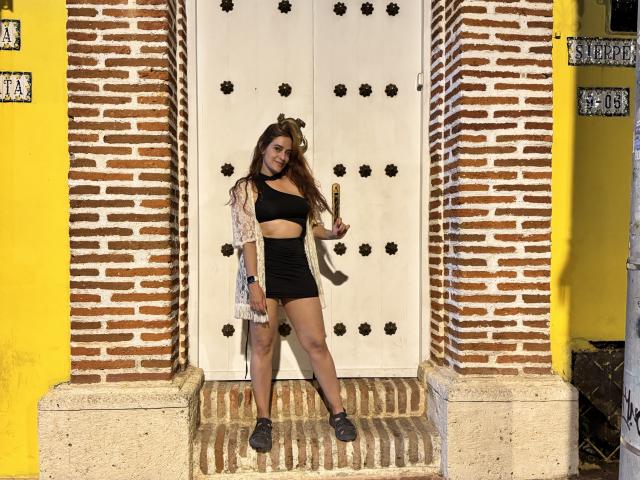 JaneRoa with long brown hair, wearing a black crop top, black shorts, and a light jacket, standing in front of a white door with black floral designs, and yellow walls.
