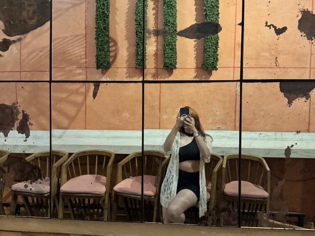 JaneRoa with tits in black bra and black panties, holding phone, mirror reflection, white lace robe, wooden chairs with pink cushions, green wall decorations, orange wall, dirty mirror.