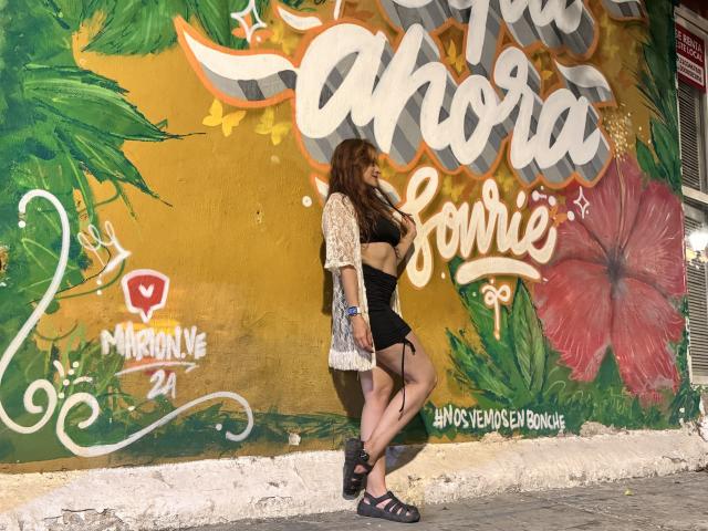 JaneRoa with brown hair, wearing a beige lace top and black shorts, standing against a colorful graffiti wall, looking to the side, with a tattoo on her right arm.