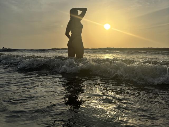 JaneRoa stands in the ocean at sunset, silhouetted against the sun, with one hand on her head, water up to her thighs, and her ass and tits visible.