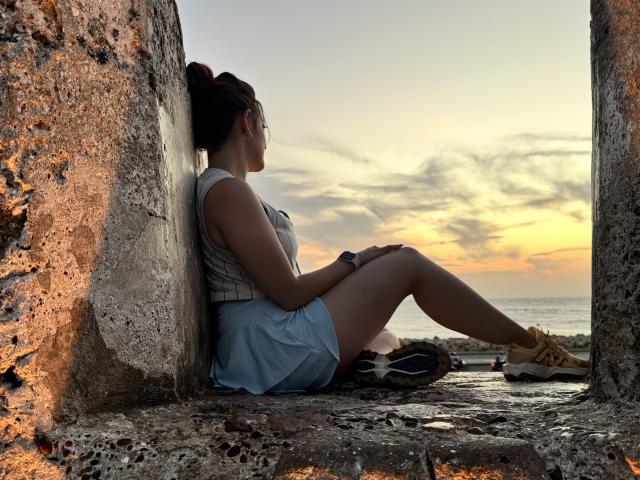 JaneRoa, with her ass resting on a rocky ledge, wears a sleeveless top and blue skirt, sitting with legs up, sneakers on, sunset in background, dark hair in a bun.