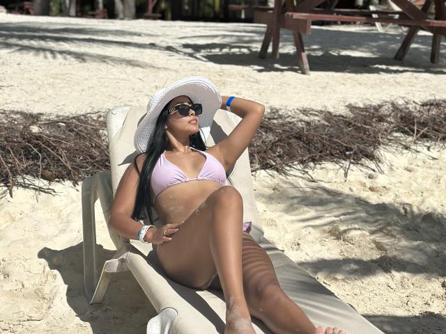 VanessaNova, de piel oscura, con cabello largo y negro, vestida con un bikini rosa, un sombrero de sol blanco, gafas de sol y una pulsera azul, descansando en una playa arenosa, con las piernas cruzadas y arena en sus muslos..
