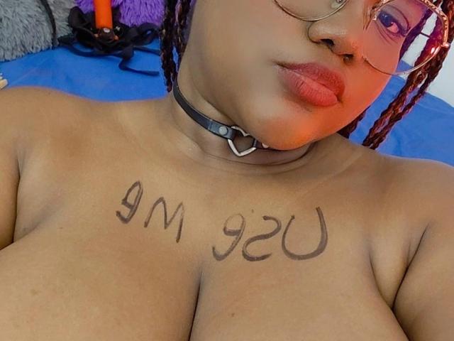 RoseBeckers, con cabello oscuro trenzado, usando gafas y un collar negro con un corazón, tiene tatuado "9M 92" en sus pechos. Está sin sujetador, mostrando sus grandes senos..