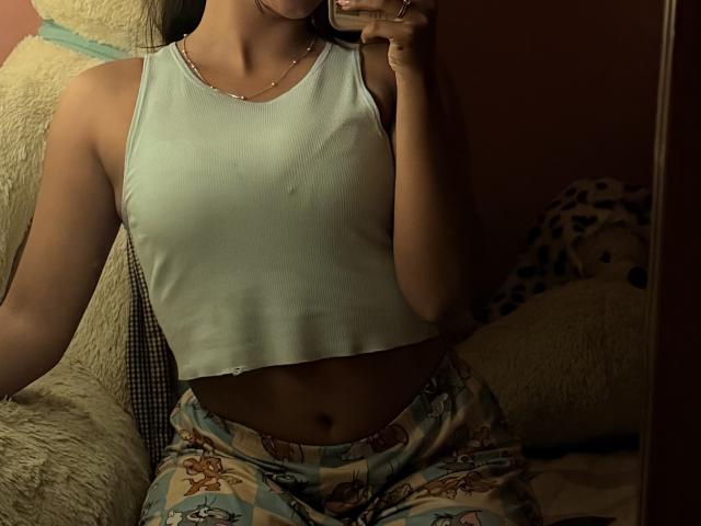 SarahPrincess with small tits, wearing a light green crop top and patterned pajama pants, holding a phone in a dimly lit room, showing her flat stomach.