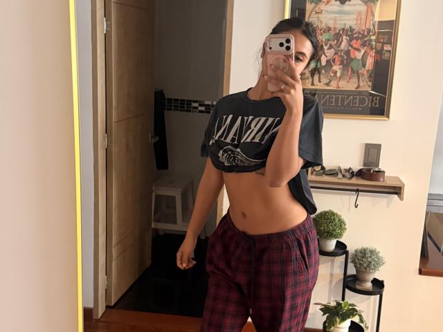 SarahPrincess with dark hair in a ponytail, wearing a black crop tee, red plaid pajama pants, showing her bare midriff and holding a pink iPhone.