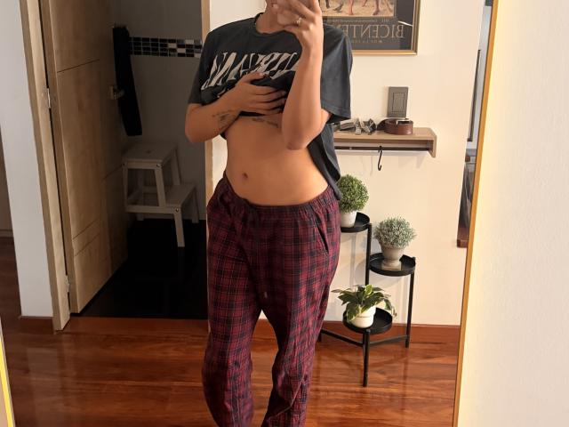 SarahPrincess in red plaid pajama bottoms, lifting a gray "Wisconsin" shirt to show a flat, tattooed midriff, standing in a mirror selfie.