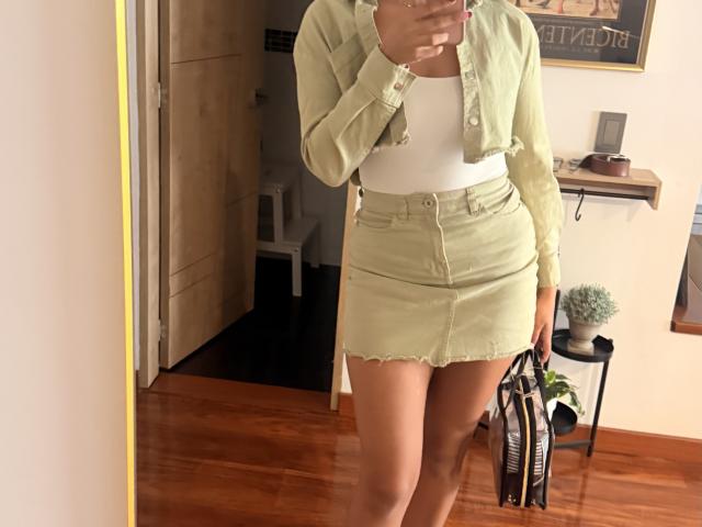 SarahPrincess wearing a green button-up crop jacket, white tank top, and high-waisted green denim skirt, holding a black and white bag, showing her ass in a mirror selfie.