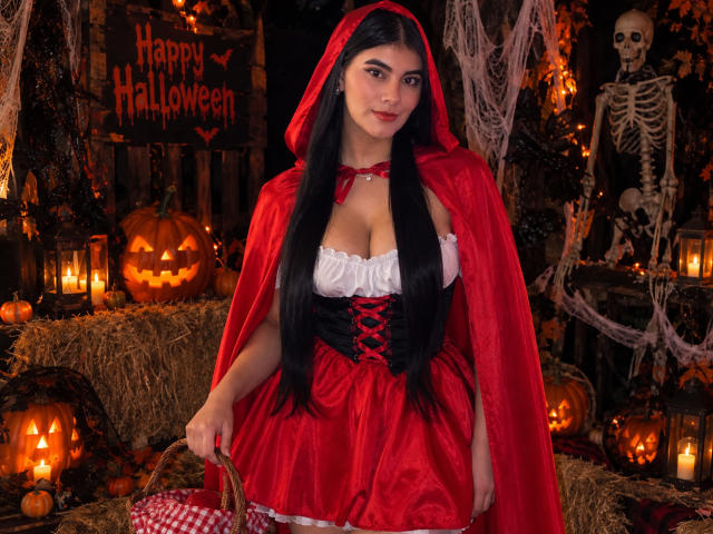 AlaraKey with long black hair, red hooded cape, and red dress with white corset, showing her big boobs, standing in a Halloween setup with jack-o'-lanterns and candles.