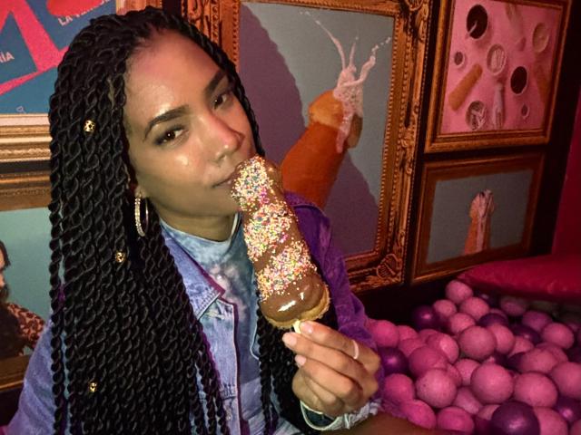 NatLewis with long black braids, eating a chocolate ice cream topped with colorful sprinkles, wearing a denim shirt and hoop earrings, in a pink and gold room.