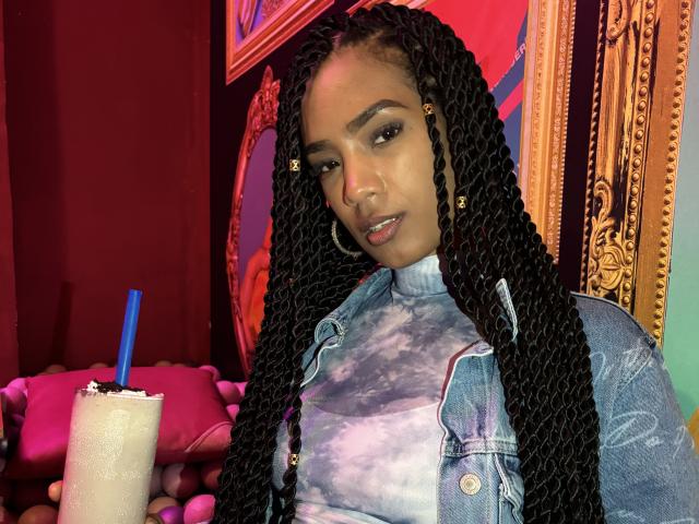 NatLewis with long black braids, wearing a tie-dye top and denim jacket, holding a drink with a blue straw, has a cute nose and a subtle smile.
