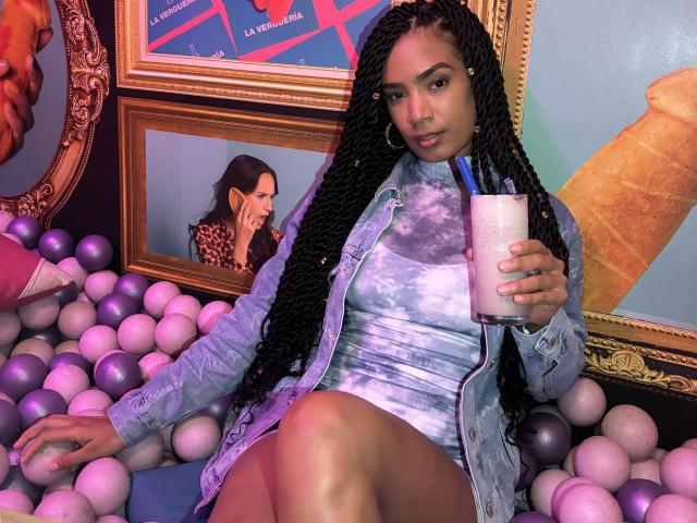 NatLewis with long braids, wearing a denim shirt and tie-dye dress, sitting in a ball pit, holding a pink cup with pens, showing her smooth ass and calm expression.
