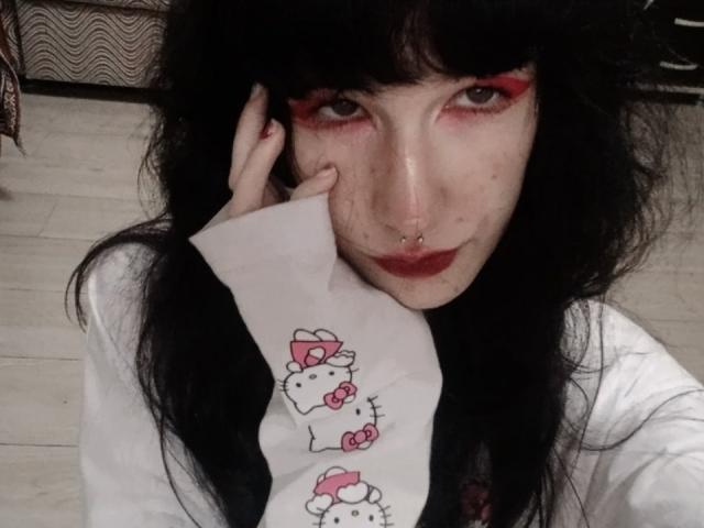 Vinita Bartolomei, con capelli neri, trucco occhi rosso, piercing al naso e una camicia bianca con la stampa di Hello Kitty, che tocca il viso con un foglio di carta, con la pelle pallida e il rossetto rosso..