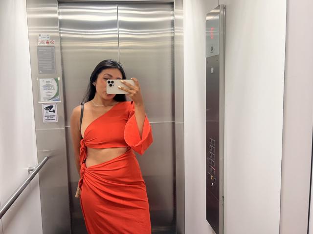 AmeliRoux in a shiny elevator, wearing a red one-shoulder dress with a cutout, holding a phone to her face, long black hair, small tits, slim, sexy, reflecting mirror.