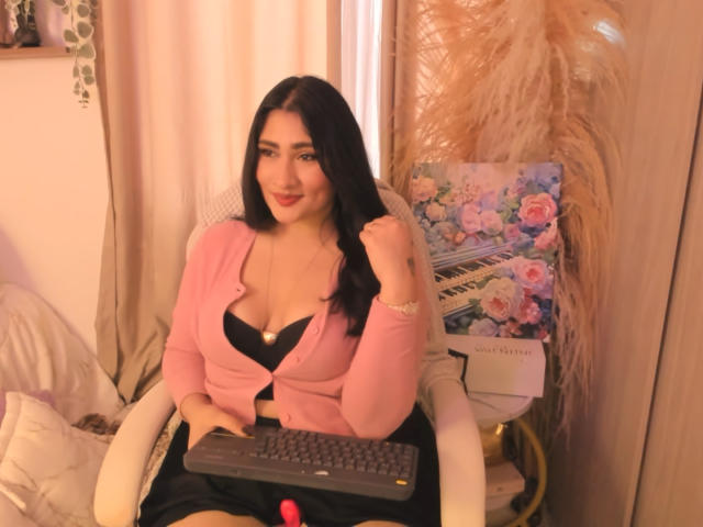 ElyRoss, avec ses cheveux noirs, son cardigan rose, son soutien-gorge noir et sa poitrine dégagée, assise sur une chaise, tenant un clavier, souriante, avec des plumes roses duveteuses et une décoration florale derrière elle..
