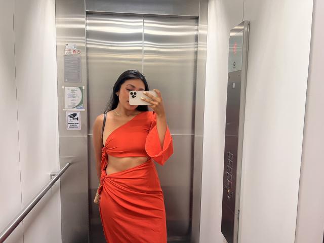 AmeliRoux with long black hair, wearing an orange, one-shoulder, cut-out dress, taking a mirror selfie in a silver elevator, showcasing her ass and flat stomach.