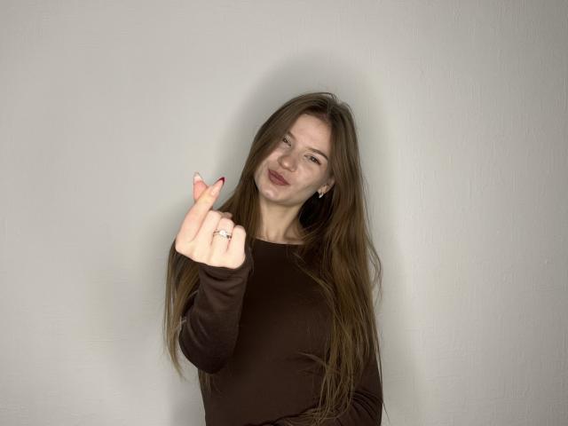 JustineHasenfratz with long brown hair, brown long-sleeve shirt, pointing with red-manicured fingers, wearing a ring, looking slightly annoyed.
