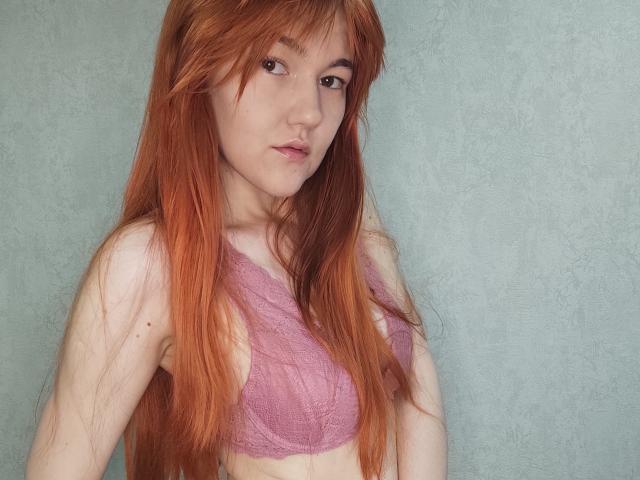 SheritaChieng has long red hair, wearing a pink lace bralette, showing small tits and a slim, pale body, with a neutral expression and green eyes, against a plain background.