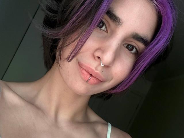 TameikaMacclellan has purple and black hair, a nose ring, and a lip ring, with pale skin and brown eyes, wearing a white strap, looking at the camera.