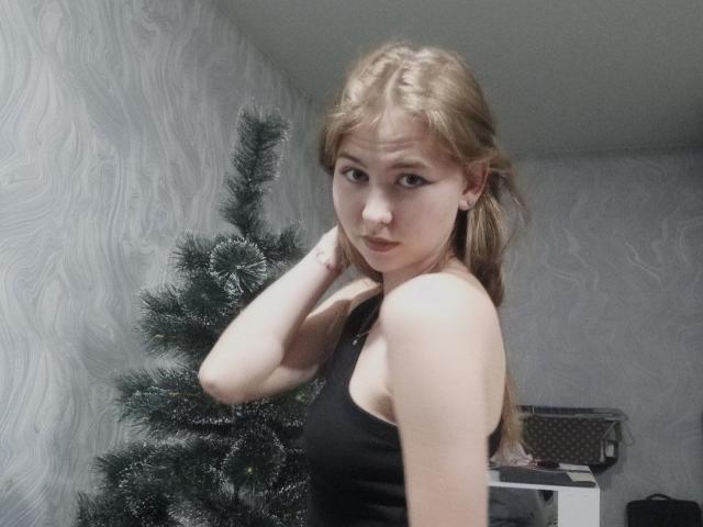 MargretWarriner with blonde hair, small tits, black tank top, hand on neck, standing next to a Christmas tree, looking at camera, in a gray-walled room.