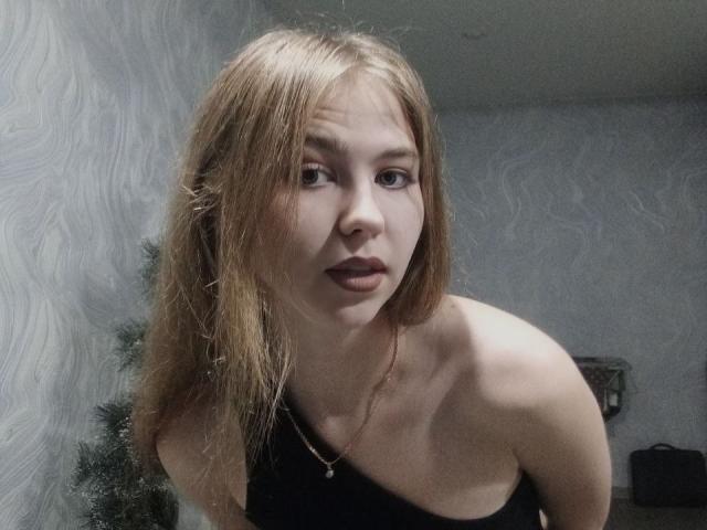 MargretWarriner with wet blonde hair, wearing a black top, has small tits and a slim figure, looking at the camera with dark lipstick, in a dimly lit room.