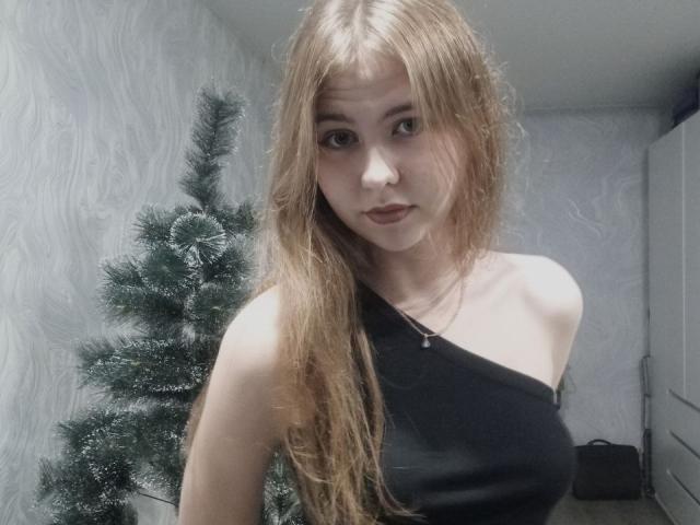 MargretWarriner with long red hair, black one-shoulder top, small tits, pale skin, and a neutral expression, standing next to a green, snow-dusted Christmas tree.