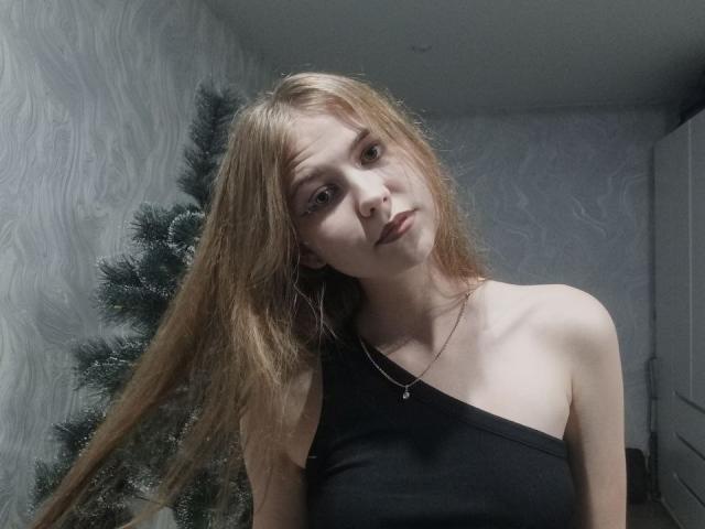 MargretWarriner with long blonde hair, wearing a black one-shoulder top, slight smirk, fair skin, necklace, in a room with a Christmas tree.