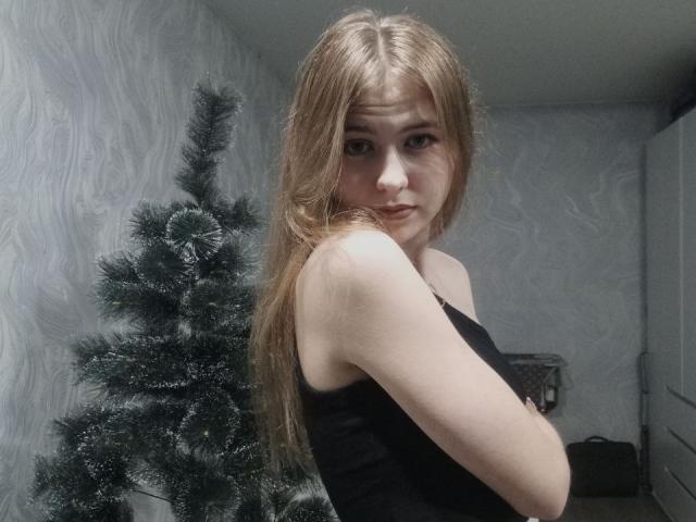MargretWarriner with long red hair, black tank top, looking over shoulder, standing beside a small, green, fake Christmas tree. Her tits are visible, she has a slightly confused expression.