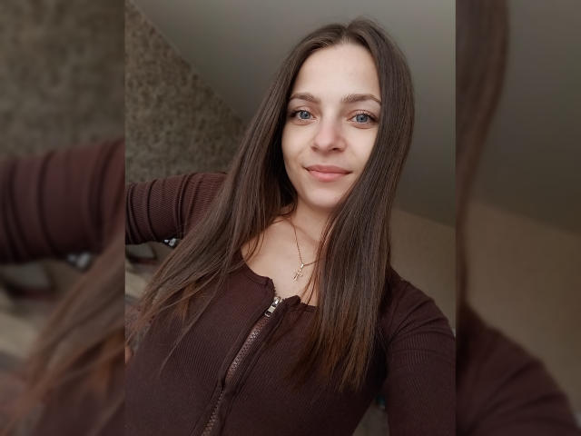 SaraUili with long brown hair, blue eyes, and a slight smile, wearing a brown zip-up top, standing in front of a mirror, reflecting her upper body.