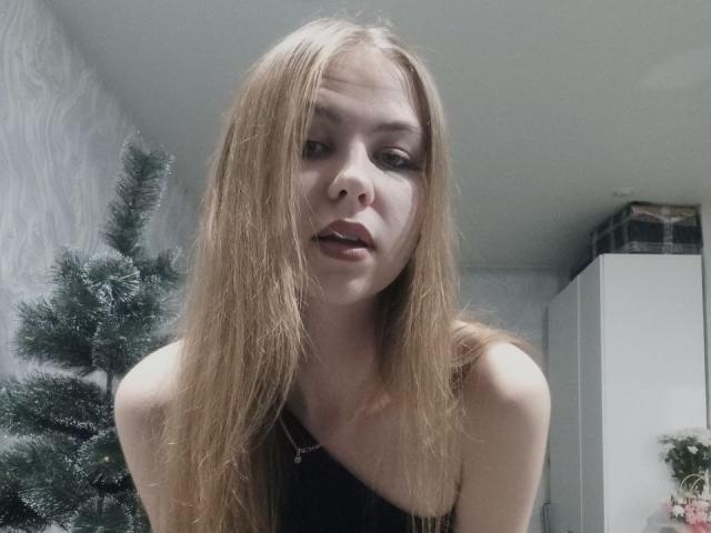 MargretWarriner with long, dirty blonde hair, wearing a black top, looking down with slightly parted lips, showing her bare shoulders and upper chest. A small Christmas tree is behind her.