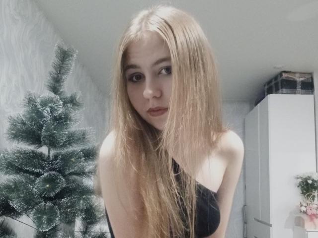 MargretWarriner with long blonde hair, small tits, wearing a black strapless top, standing in front of a green, snow-dusted Christmas tree, looking at the camera with a serious expression.