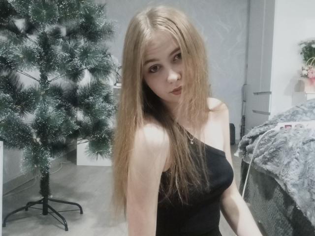 MargretWarriner with long blonde hair, wearing a black one-shoulder top, showing small tits, standing in front of a green Christmas tree, looking at the camera with a neutral expression.