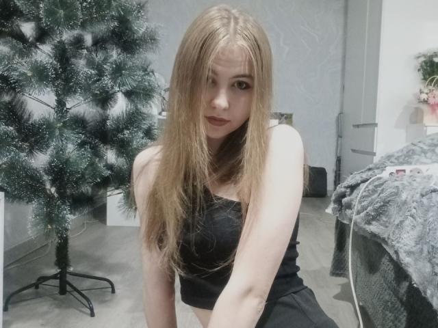 MargretWarriner with long blonde hair, wearing a black strapless top, sitting in front of a small Christmas tree, looking at the camera with a slightly serious expression.