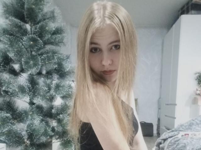 MargretWarriner with long blonde hair, black top, looking at camera, standing in front of a green, frosty Christmas tree, slight pout, fair skin, cute, attractive.