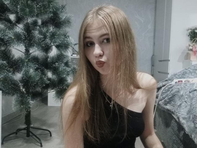 MargretWarriner with long brown hair, black strapless top, and pouty lips, standing next to a small Christmas tree, wearing a necklace with a cross. Boobs are partially visible.