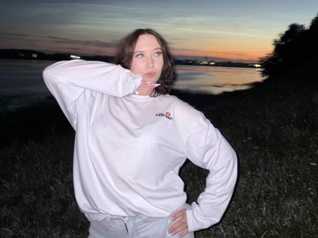 LasonyaKaehler with wavy brown hair, white Adidas sweatshirt, and hand on hip, pouting with finger to cheek, standing on grassy edge at dusk.