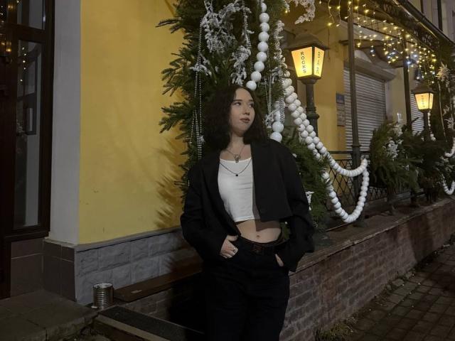 LasonyaKaehler stands against a yellow wall, wearing a black blazer, white crop top, and black pants, with hands in pockets, looking up, dark curly hair, small tits, medium ass.