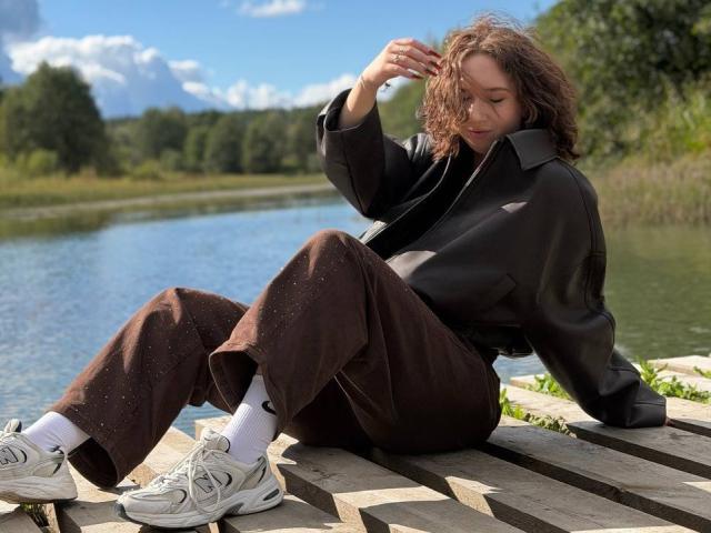 LasonyaKaehler with curly brown hair, black jacket, brown pants, white socks, and beige sneakers, sitting on wooden planks by a lake, adjusting her hair.
