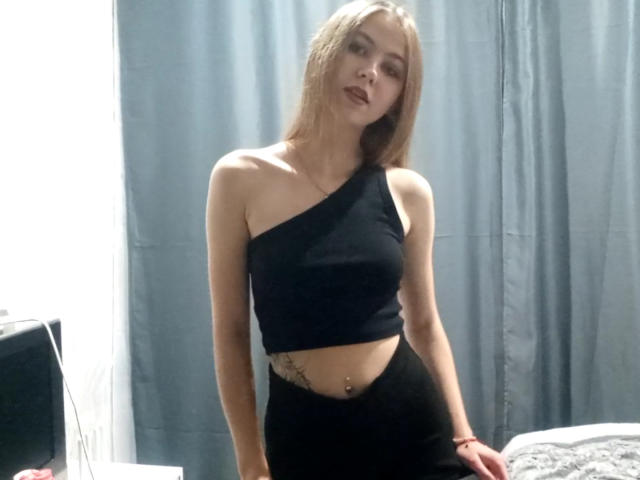 MargretWarriner with blonde hair, one-shoulder black crop top, black pants, belly button piercing, tattoo under left breast, standing in bedroom, slightly leaning forward.