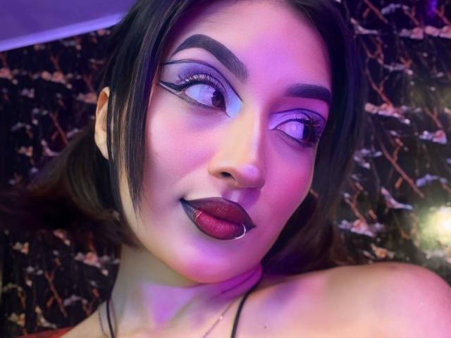 LunaixMoon with dark, shiny lipstick, thick black eyeliner, and a silver lip piercing, looking to the side with purple and blue lighting on her face. She has black hair.
