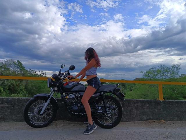LunaTomson, com cabelo ombré vermelho, vestindo uma blusa cinza e shorts pretos, sentada em uma motocicleta preta, com as mãos nos guidões, em um cenário ensolarado com um céu nublado..