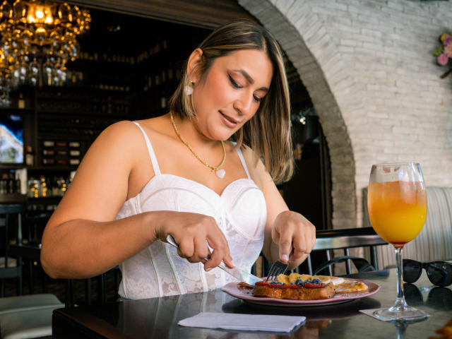 EmaFoxy con caschetto castano, bustino di pizzo bianco, seno di media grandezza, che mangia da un piatto, tenendo un coltello e una forchetta, bevanda arancione davanti, lampadario, parete di mattoni bianchi, collana..
