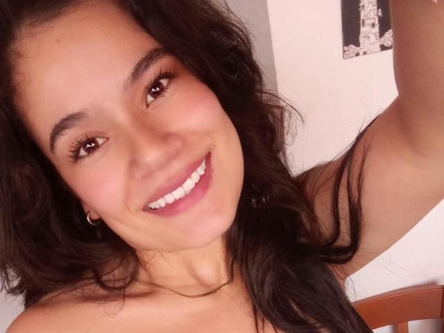 IsabellaVoss with wavy black hair, brown eyes, and a cute smile, showing white teeth, small hoop earrings, and a necklace, raising her right arm.