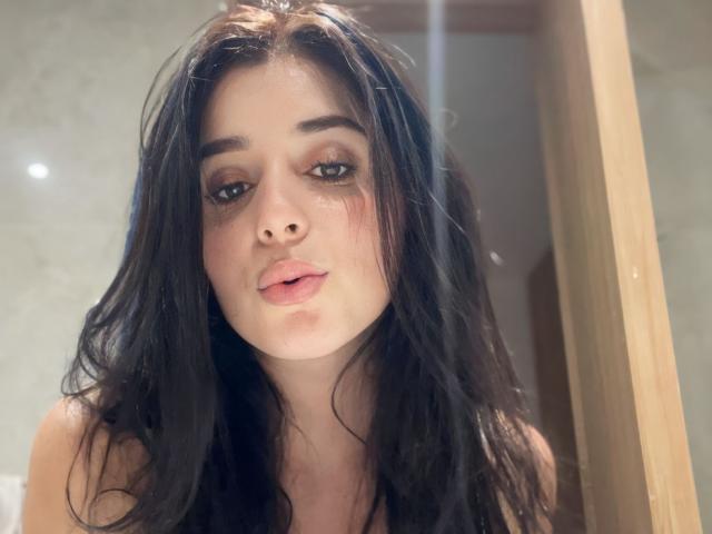 VickyMegan heeft lang, golvend zwart haar, bleke huid en volle, roze lippen. Ze staart naar een spiegel met licht open, glanzende roze lippen en donkere, smoky ogen..