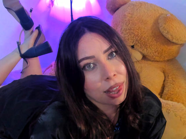 JennyLawry with dark brown hair, blue eyes, and pink lipstick, lying down, wearing black high heels, black shirt, and a cute expression, with a large brown teddy bear behind her.