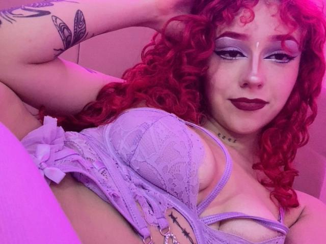 GaiaDoll com cabelo ruivo encaracolado, maquiagem roxa nas pálpebras, sutiã de renda roxo, mostrando os seios, tatuagem no braço, colar, luz roxa, batom escuro, anel no dedo, sorriso leve, pose relaxada..