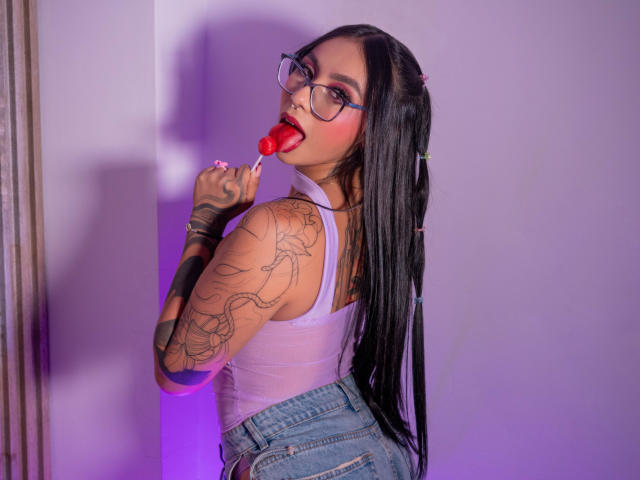 ChloeMoretti met lang zwart haar, een bril, een wit tanktopje en blauwe jeans, die een rode lolly zuigt, waarbij haar getatoeëerde armen en een aantrekkelijk achterwerk zichtbaar zijn, en die over haar schouder kijkt..