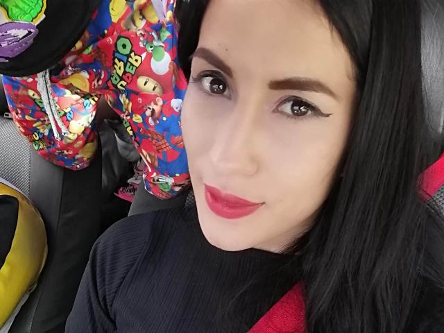 SofiaAnal with black hair, red lipstick, and winged eyeliner, wearing a black top, and colorful, cartoon-print pants, smiling at the camera, showing her cute face.